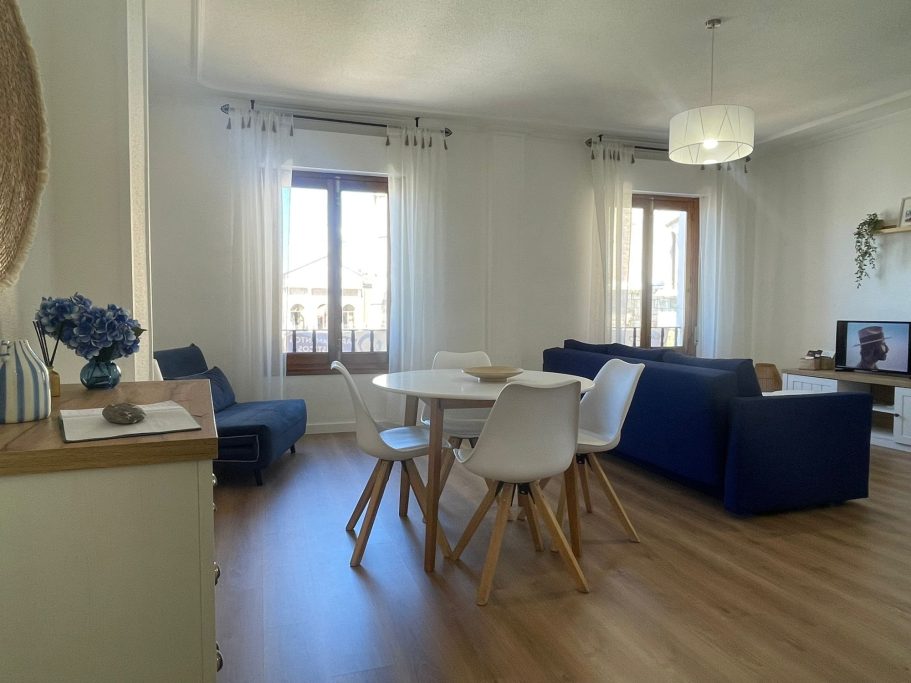 Sala de estar luminosa con sofá azul, mesa blanca y ventanas con vistas exteriores.